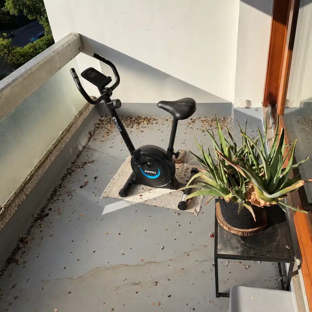 Sol de balcon avant nettoyage, avec un vélo d’appartement et une plante en pot.